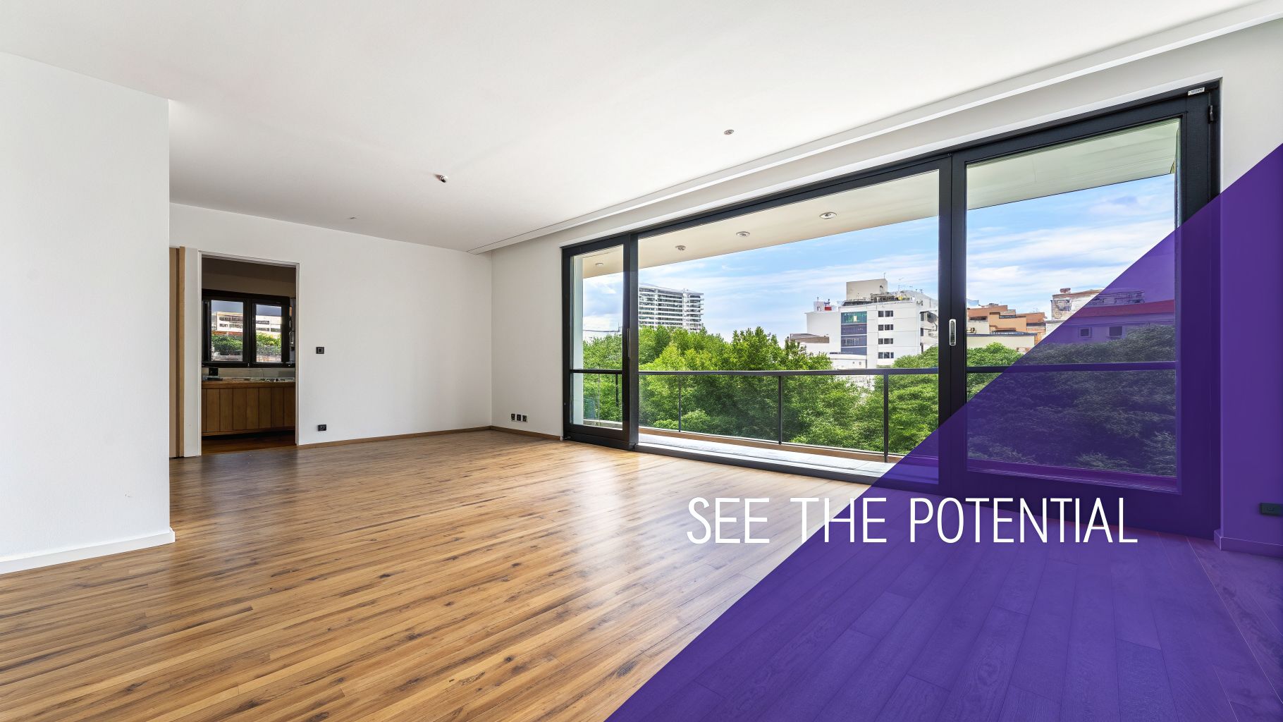 Empty modern living space with hardwood floor, large window, and city view from balcony.