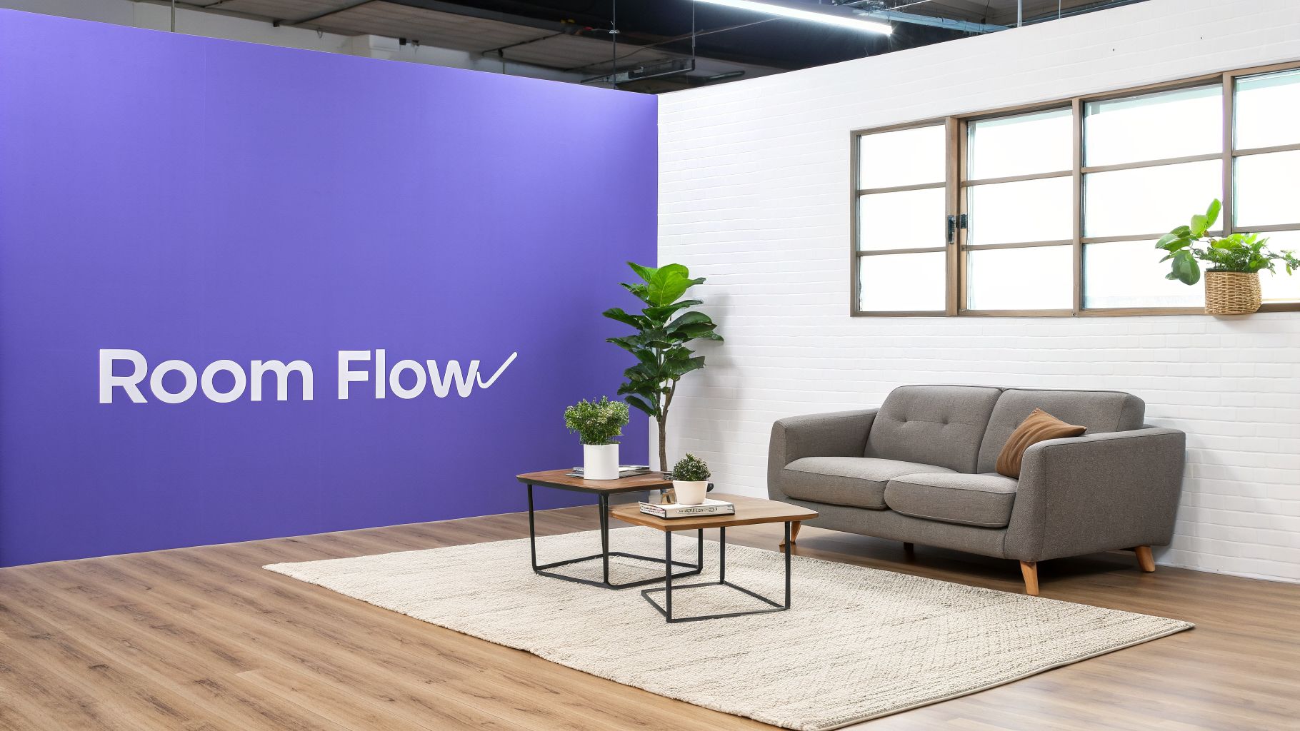 A brightly lit modern room featuring a grey sofa, coffee tables, plants, and a purple Room Flow branded wall.