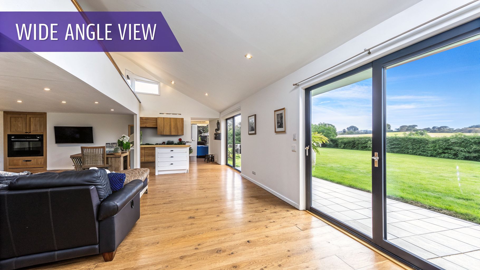 A bright, open-plan living room and kitchen with wooden floors, a large sofa, and patio doors leading to a green garden.